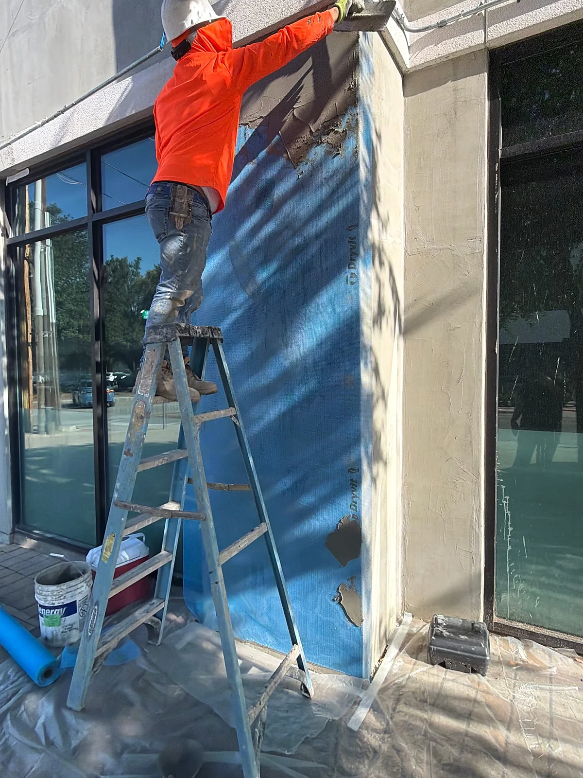 Worker applying gray material over facade mesh beside ground-floor glazing in Dallas.